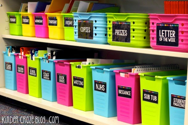 bright pink, green, blue and yellow bins labeled for school supply storage on a classroom shelf
