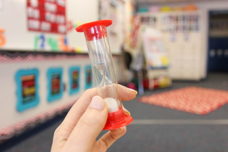 teacher holding a small sand timer in a kindergarten classroom. Classroom background with an easily, framed classroom rules, whiteboard and student rug are blurred in background.