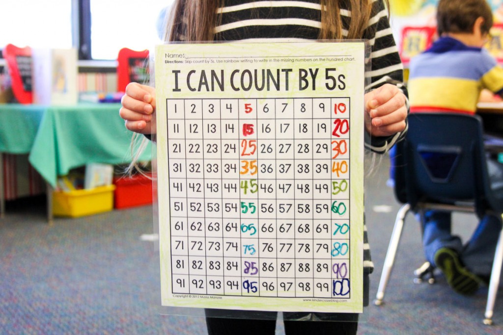kindergarten student in classroom holding up completed skip counting chart that with colorful rainbow numbers