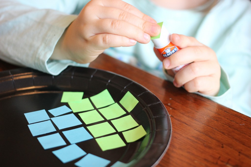 child gluing blue and green squares onto a black paper plate to create a simple earth day craft for kids