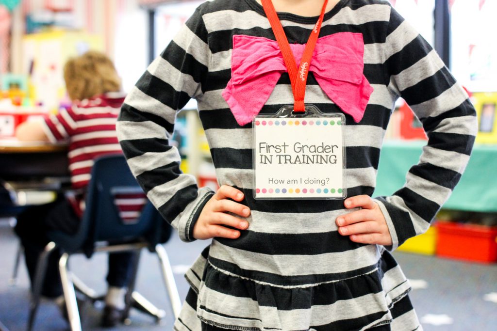 Kindergarten student standing in a classroom wearing a First Grader in Training Badge around her neck