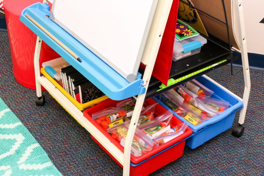 large storage bins under a classroom easel. Each bin is filled with clear plastic bags containing school supplies for each student in the class that are clearly labeled.