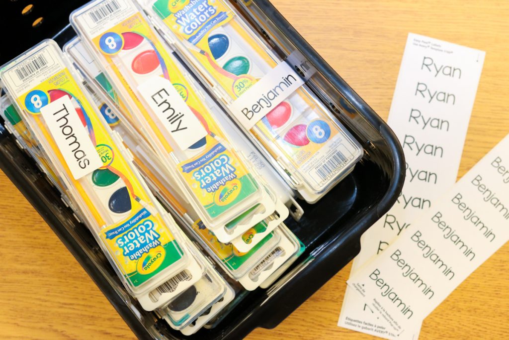 black plastic classroom bin containing boxes of watercolor paints lableed with student names. Two sheets of printed labels lay beside the bin read to peel and stick for the next student.