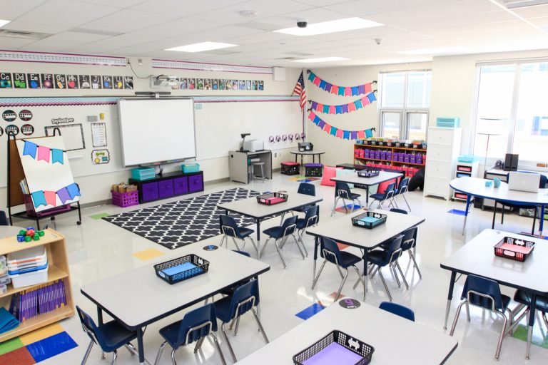 bright first grade classroom with table seating, white walls and large black and white classroom rug