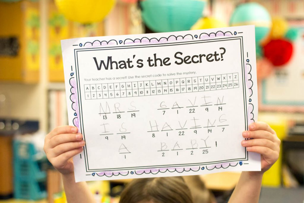 student in classroom holding up a completed secret code worksheet that says "My Teacher is Having a Baby" for a teacher pregnancy reveal