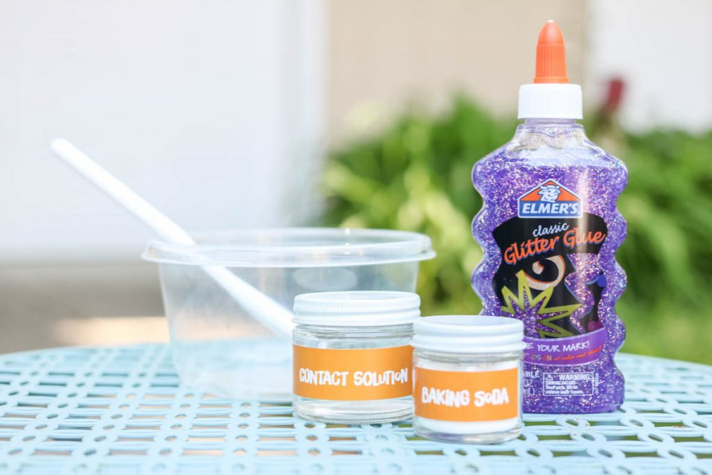 3 simple ingredients to make slime at home displayed on a table outside: baking soda, contact solution and elmers glitter glue