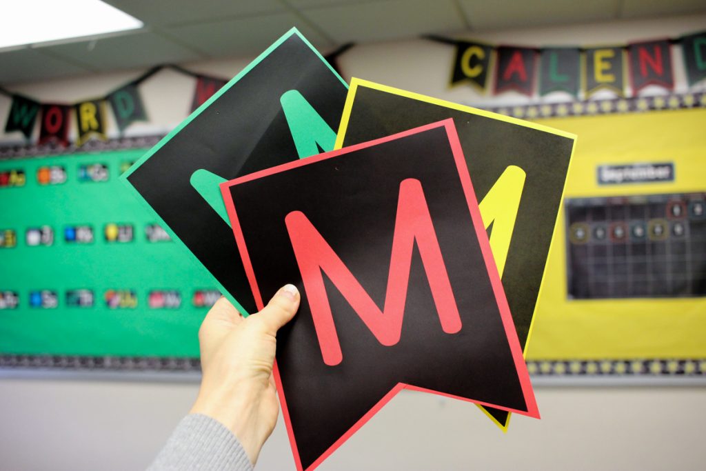teacher holding up bulletin board pennant letters in first grade classroom printed on red, green, and yellow colored paper