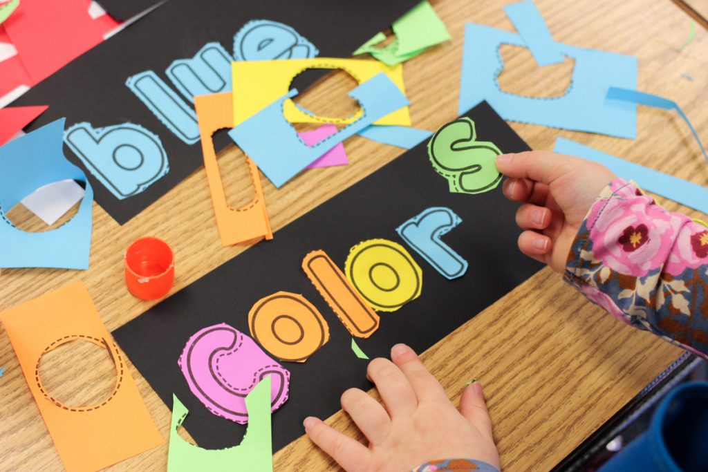 child unscrambling cut out letters and gluing them to spell "colors" on black piece of paper