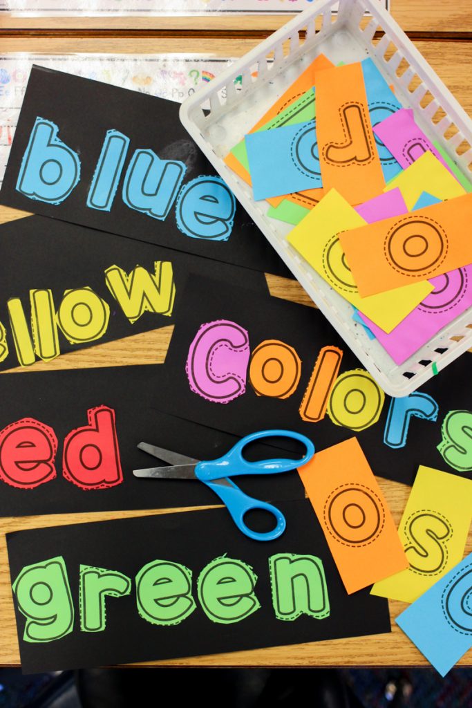 black color word book pages with colored letters glued on to spell blue, yellow, red, green, and colors laying on kindergarten desk with a blue scissors and basket of scrambled letters placed on top