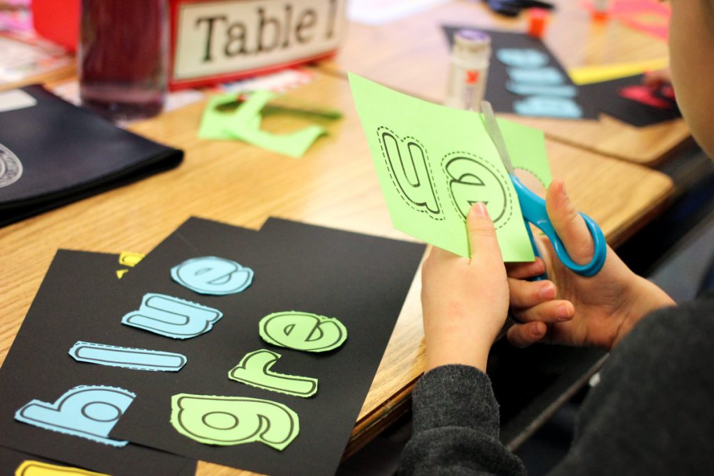 kindergarten student cutting letters to spell "green" and glue them onto a sheet of black color word worksheet