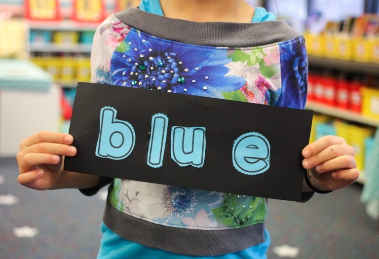 child holding up a black paper with the letters "blue" glued on it to spell color words instead of doing a worksheet
