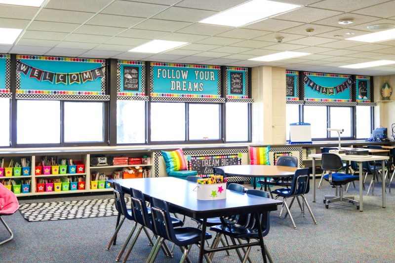 first grade classroom with brightly colored bins on library bookshelves, student desks and a black and white classroom rug