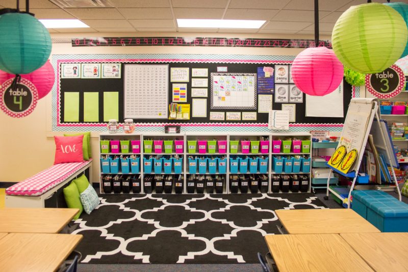 first grade classroom library with brightly colored bins on a white shelf and black carpet for students to gather on