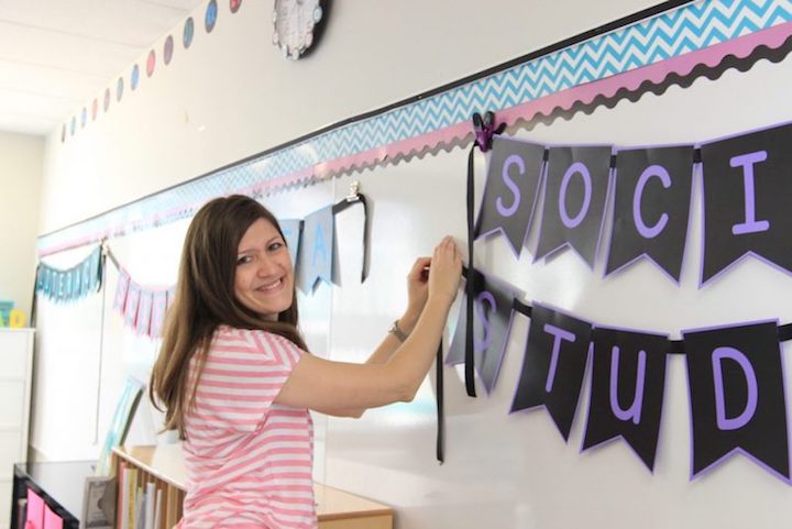 teacher setting up classroom decor with purple printed bulletin board pennant letters that spell "social studies"